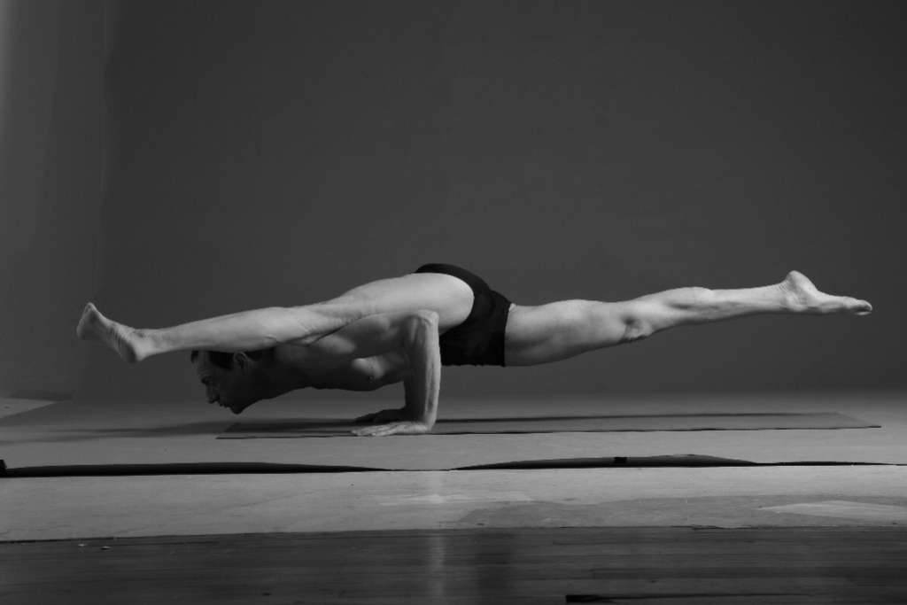 A black-and-white image of a man performing an advanced yoga pose, balancing his body parallel to the ground with legs extended in opposite directions, supported by his arms on a yoga mat in a minimalist studio setting.