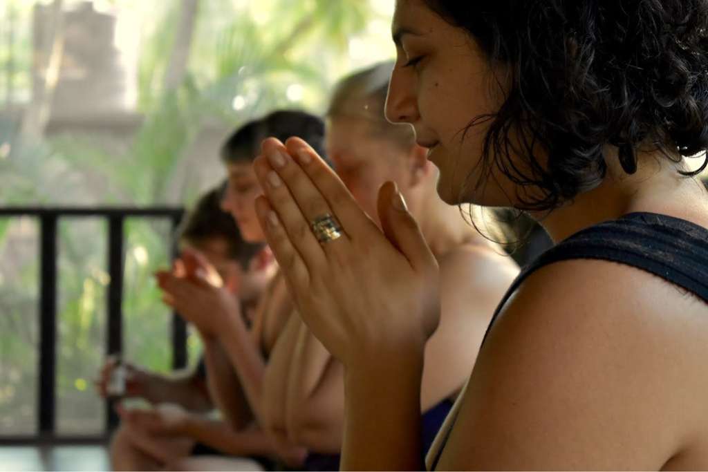 A group of people sits in a line, hands pressed together in a prayer or meditation gesture, focused and serene, with soft natural light and greenery in the background.