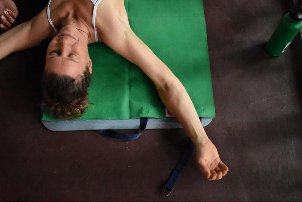 A woman lies on her back with one arm extended on a green yoga mat, practicing a relaxation pose in a calm indoor environment.