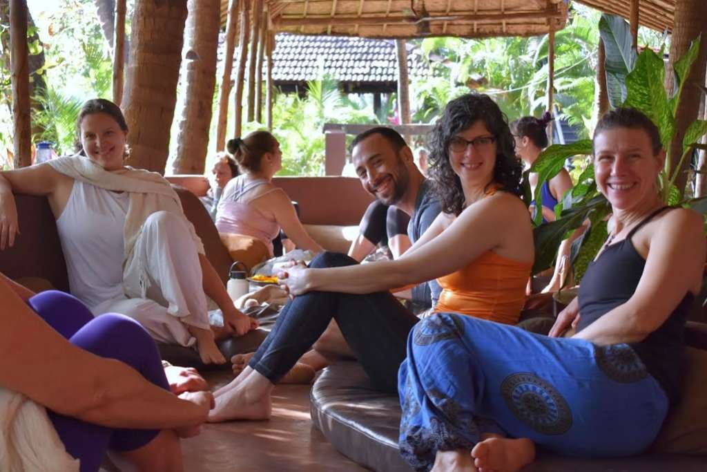 A group of people relaxes together on cushioned seating in a tropical outdoor setting, surrounded by lush greenery and natural wooden structures, enjoying a friendly and calm atmosphere.