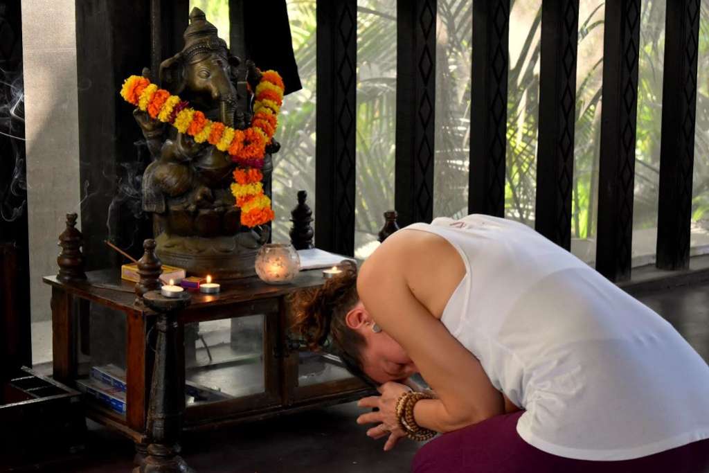 A woman bows in or meditation before a statue of Ganesha adorned with a garland of orange and yellow flowers, surrounded by lit candles and incense in a peaceful indoor setting.