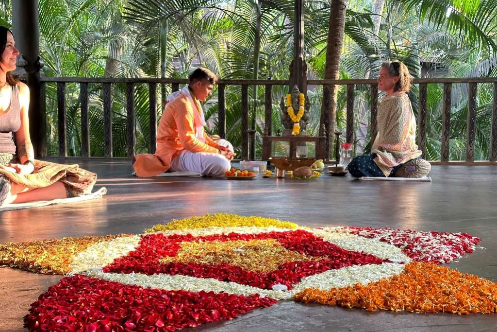 A vibrant flower mandala decorates the floor of a serene open-air pavilion where individuals sit cross-legged in meditation or prayer, with a spiritual guide conducting a ritual near a small altar adorned with flowers and offerings, surrounded by lush greenery.