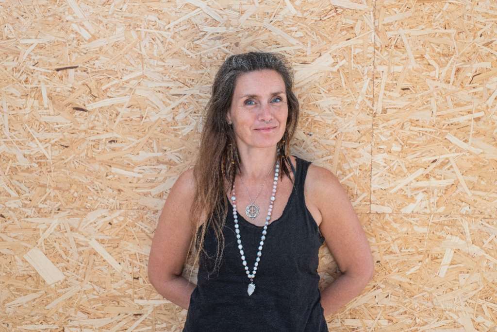 A woman with long hair wearing a black tank top and a beaded necklace stands confidently against a textured wooden background, smiling softly.