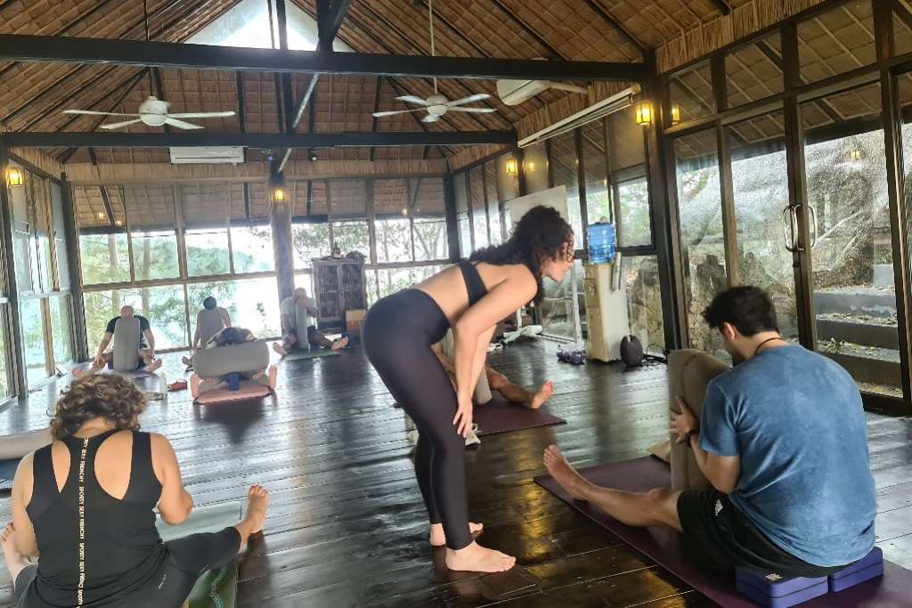 A yoga instructor providing personalized guidance to a student in a serene studio with large glass windows offering views of lush greenery. The setting features high ceilings with wooden beams, fans, and a polished wooden floor, creating an atmosphere of calm and focus. Other participants are engaged in individual stretches and poses in the background.