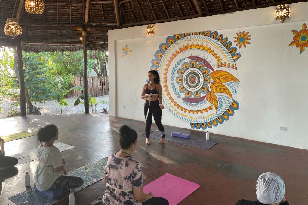 A yoga instructor stands confidently at the front of an open-air studio with a beautifully painted mandala mural on the wall behind her. Several students are seated on mats, attentively watching her. The space is warmly lit with natural light filtering through the surrounding trees, creating a serene and inspiring atmosphere.