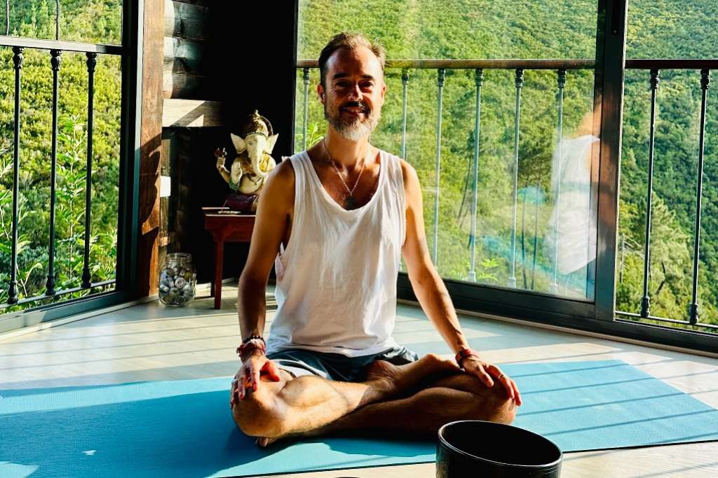 A man sits in a lotus pose on a blue yoga mat, smiling peacefully in a bright indoor space with natural light, a statue of Ganesha, and greenery visible through large windows.