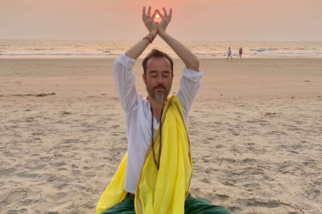 A man sits on a sandy beach in a meditative posture, wearing a white shirt and yellow shawl, with his hands raised towards the sky, framing the setting sun.