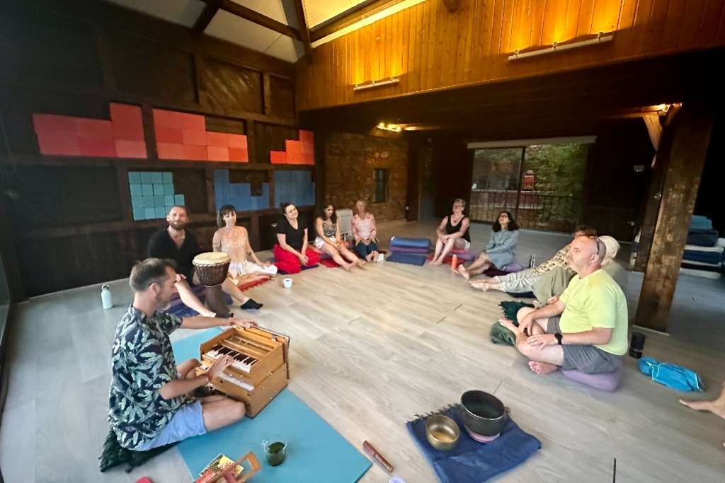 A group of people sits in a circle in a cozy indoor space, with one person playing a harmonium and others engaging in a musical or meditative session, surrounded by colorful decor and wooden accents.