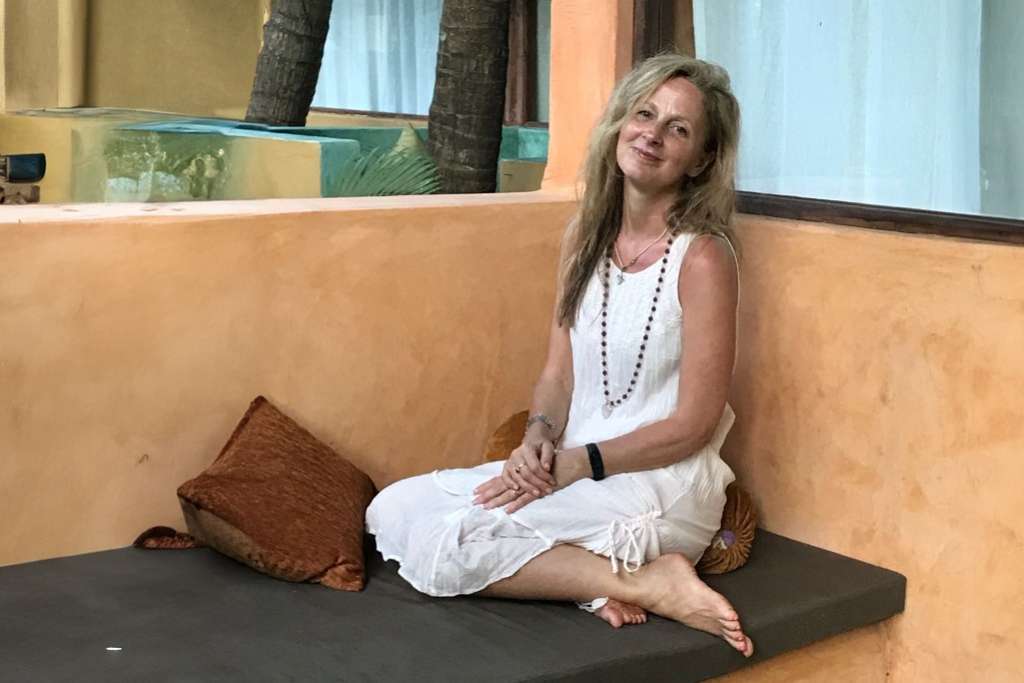 A woman in a white dress sits barefoot on a cushioned bench with a relaxed smile, surrounded by warm-toned walls and soft furnishings in a serene outdoor setting.