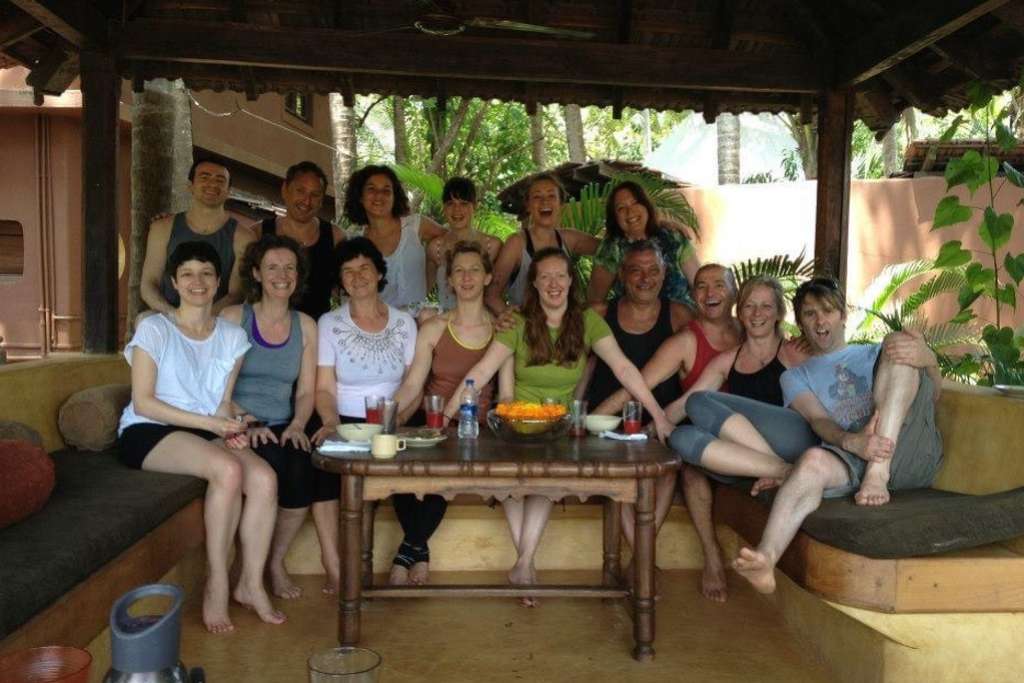 A joyful group of people gathers on cushioned seating around a wooden table in an open-air setting, smiling and enjoying a relaxed moment together surrounded by greenery.