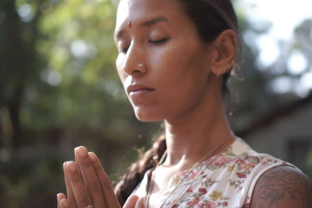 A serene woman with closed eyes and clasped hands, adorned with a bindi and floral attire, meditating outdoors.