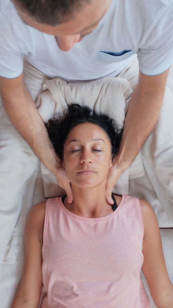 A woman in a pink tank top lies peacefully with her eyes closed while a practitioner provides a gentle neck massage or adjustment, creating a calm and therapeutic setting.