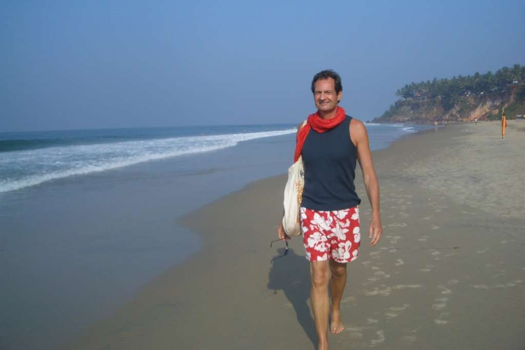 A man walks along a sandy beach under a clear blue sky, wearing a sleeveless navy top, red floral shorts, and a red scarf, carrying a bag and sunglasses with a relaxed smile.