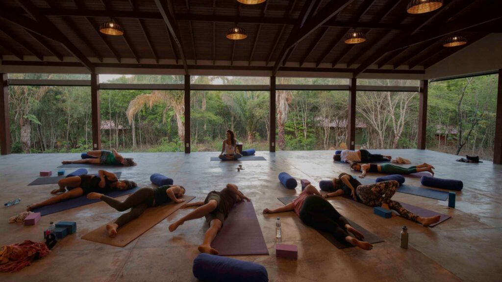 A group of yoga practitioners lies in restorative poses on mats with bolsters and props in a serene, open-air pavilion surrounded by lush greenery, with a yoga instructor seated at the center of the space.