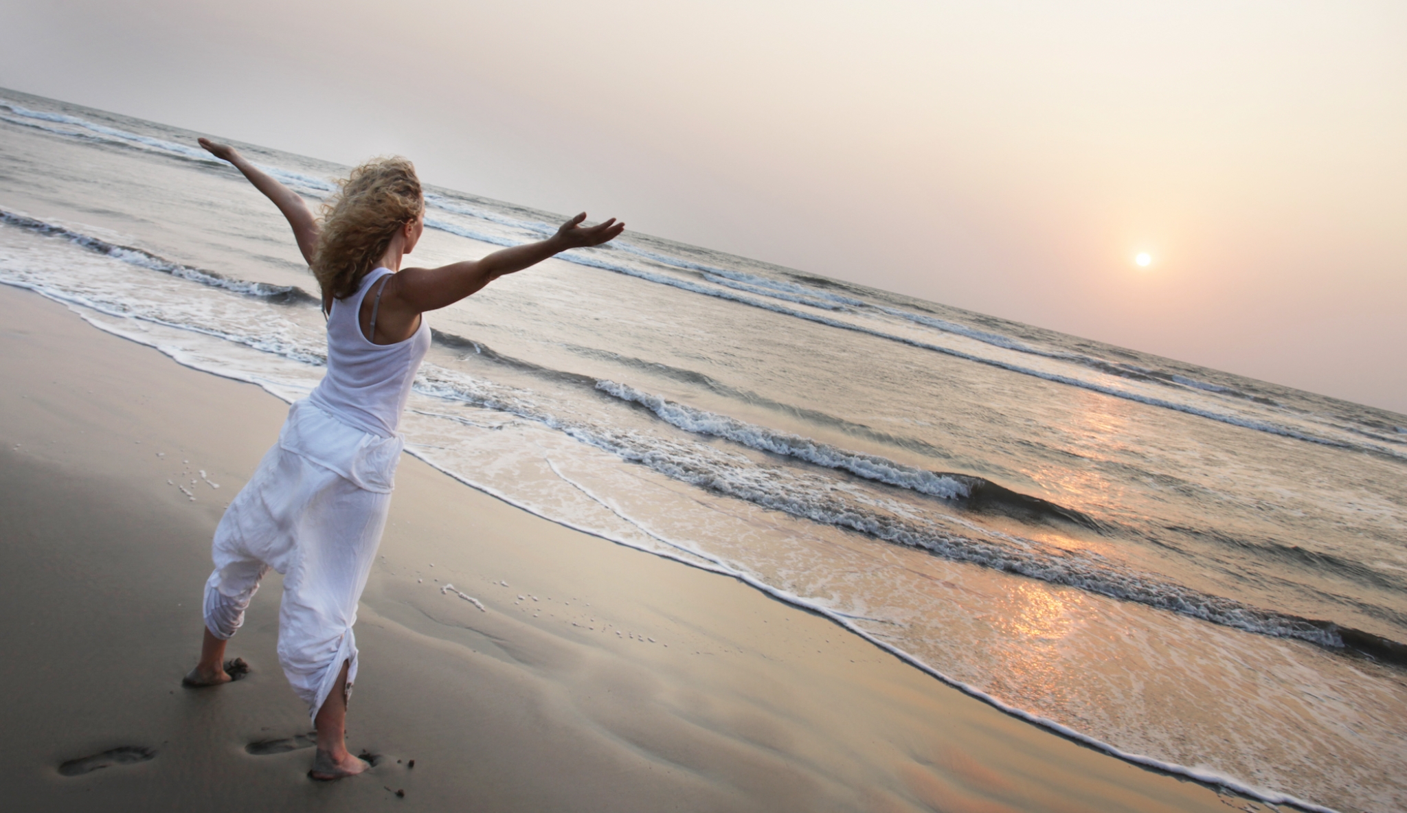 A woman wearing white clothes stands barefoot on a sandy beach with her arms outstretched, facing the ocean waves and the rising or setting sun.