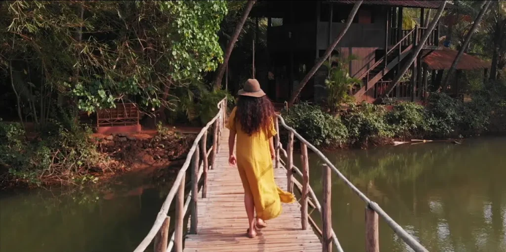 A woman in a flowing yellow dress and a wide-brimmed hat walks across a rustic wooden bridge surrounded by lush greenery, heading towards a wooden cabin nestled among trees by the water.