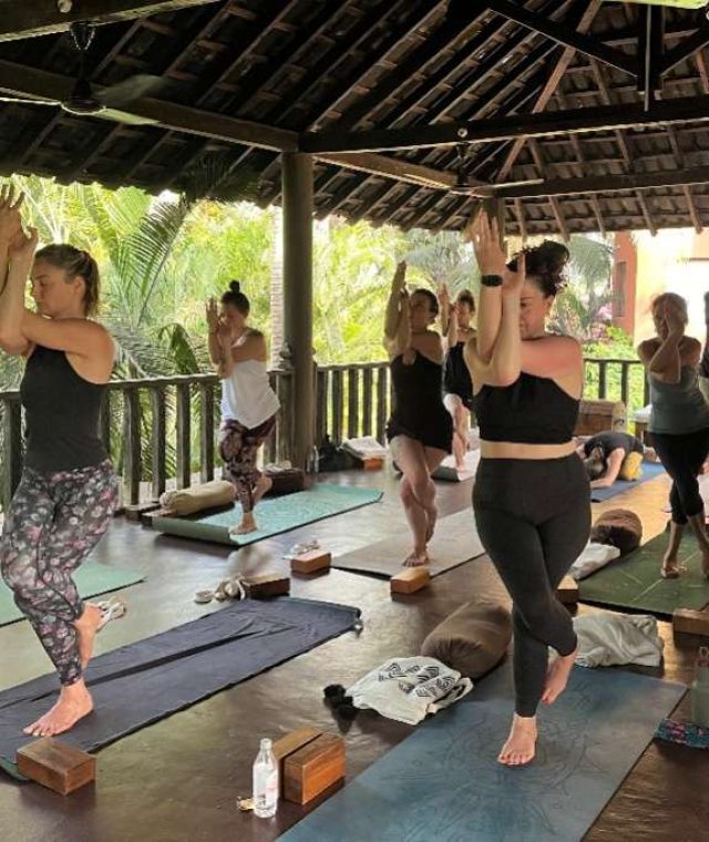 A group of people practices yoga in a spacious open-air pavilion surrounded by lush greenery, performing balancing poses with focus and concentration.